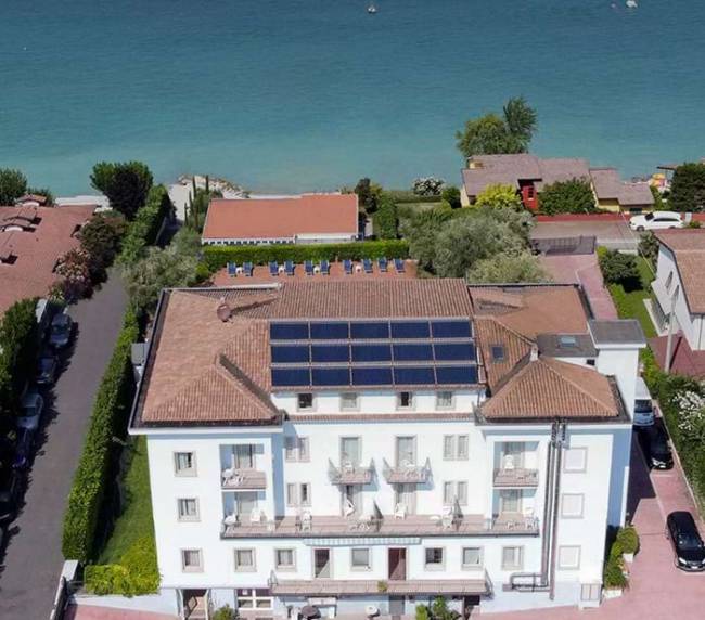 Aerial view of a white, multi-story building by the sea with solar panels on its terracotta roof and balconies facing the water.