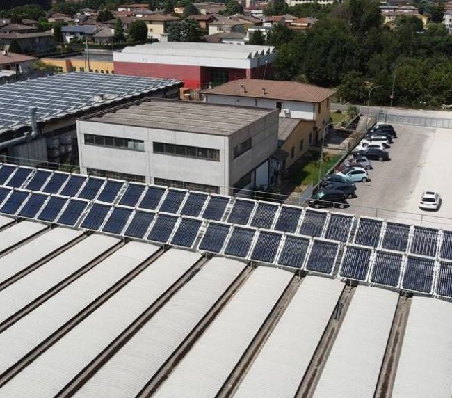 Line of solar panels installed on the roof of a large industrial building, with a parking lot and neighborhood beyond.