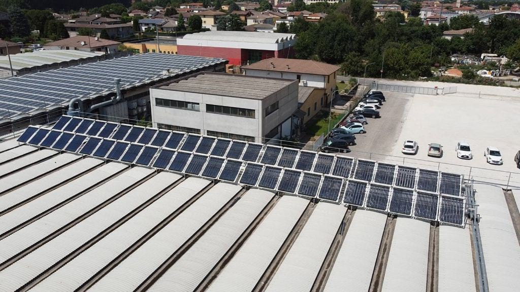 Verona-impianto-solare-termico - Pleion Line of solar panels installed on the roof of a large industrial building, with a parking lot and neighborhood beyond.