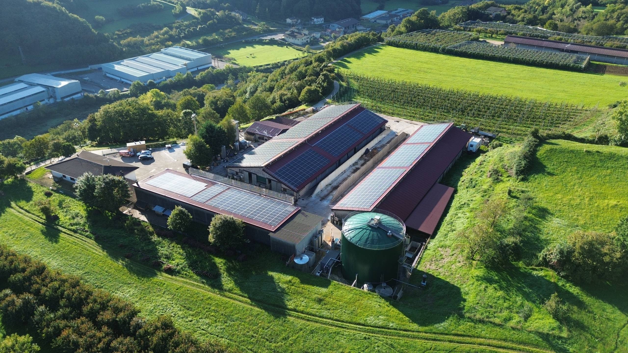 DCIM100MEDIADJI_0184JPG - Pleion Aerial view of an industrial complex with solar panels covering long roofs, surrounded by green fields and trees.