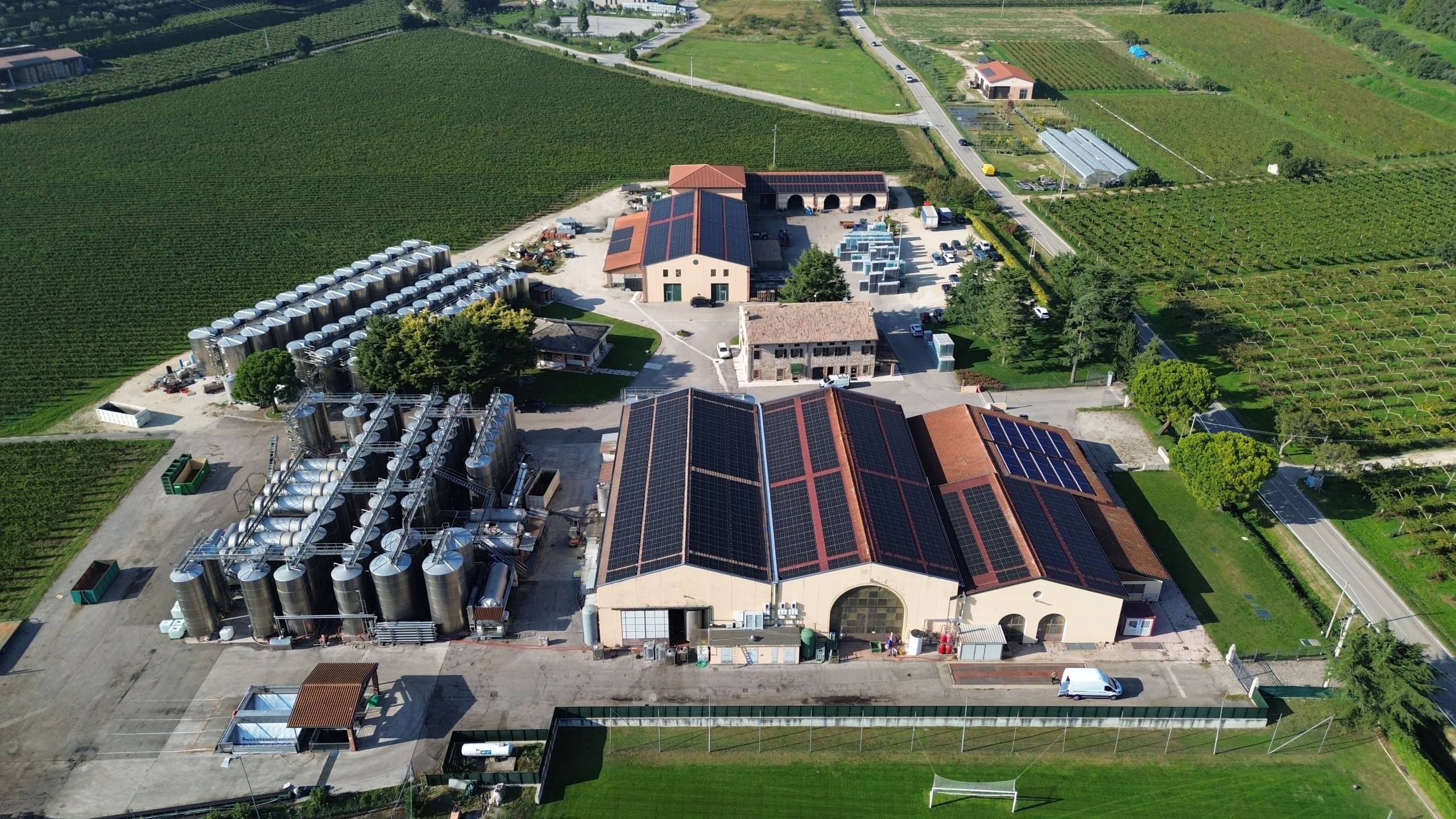 DCIM100MEDIADJI_0272JPG - Pleion Aerial view of a farm complex with solar panels on rooftops, many silos on the left, and surrounding vineyards and green fields nearby.