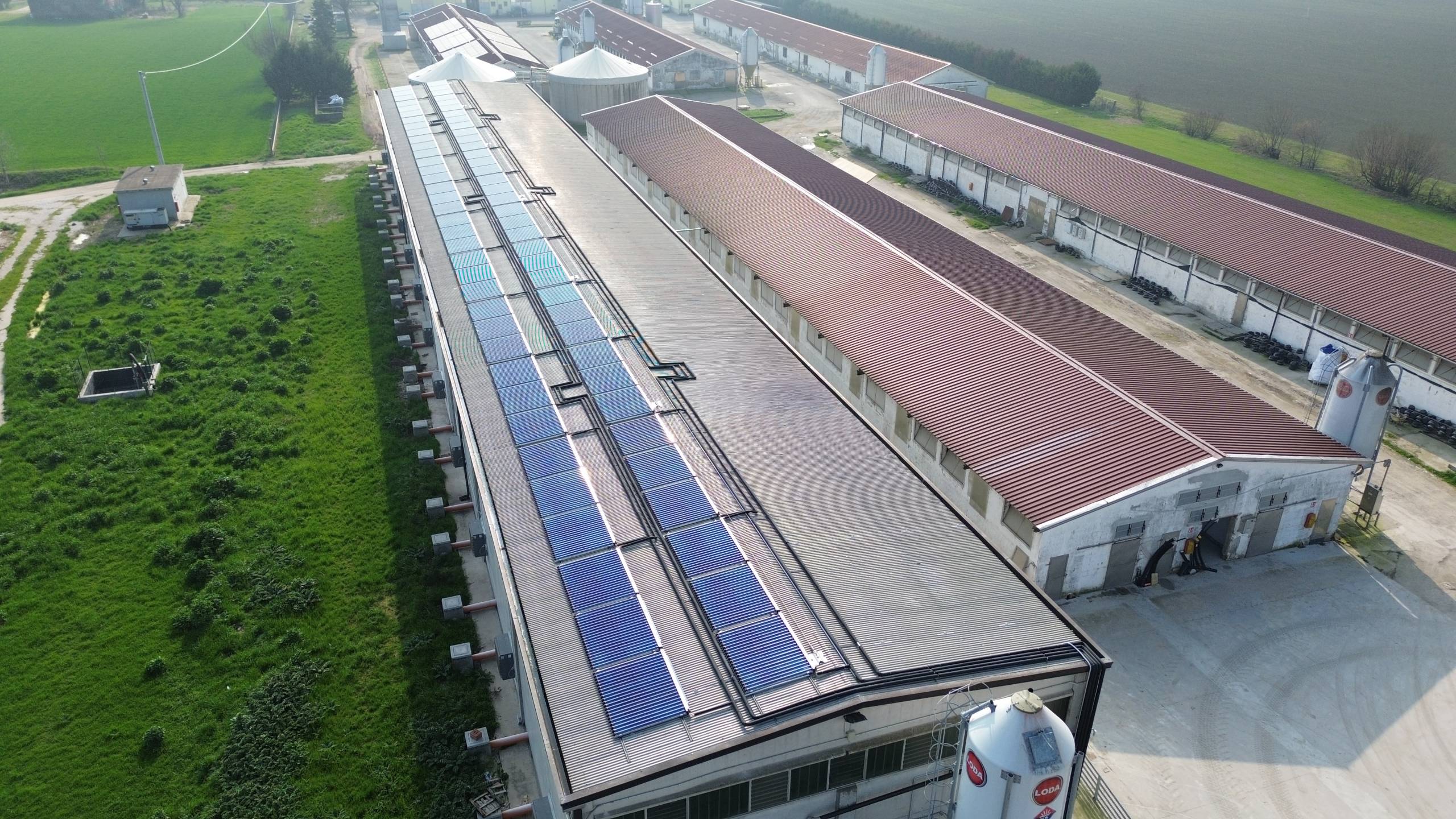 DCIM100MEDIADJI_0445JPG - Pleion Aerial view of a long industrial barn complex with rows of solar panels on the roof and green fields on one side and farmland on the other side.