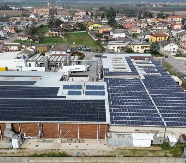 Aerial view of an industrial facility with large solar panels covering the roof, loading bays and delivery trucks nearby, and a residential town beyond.
