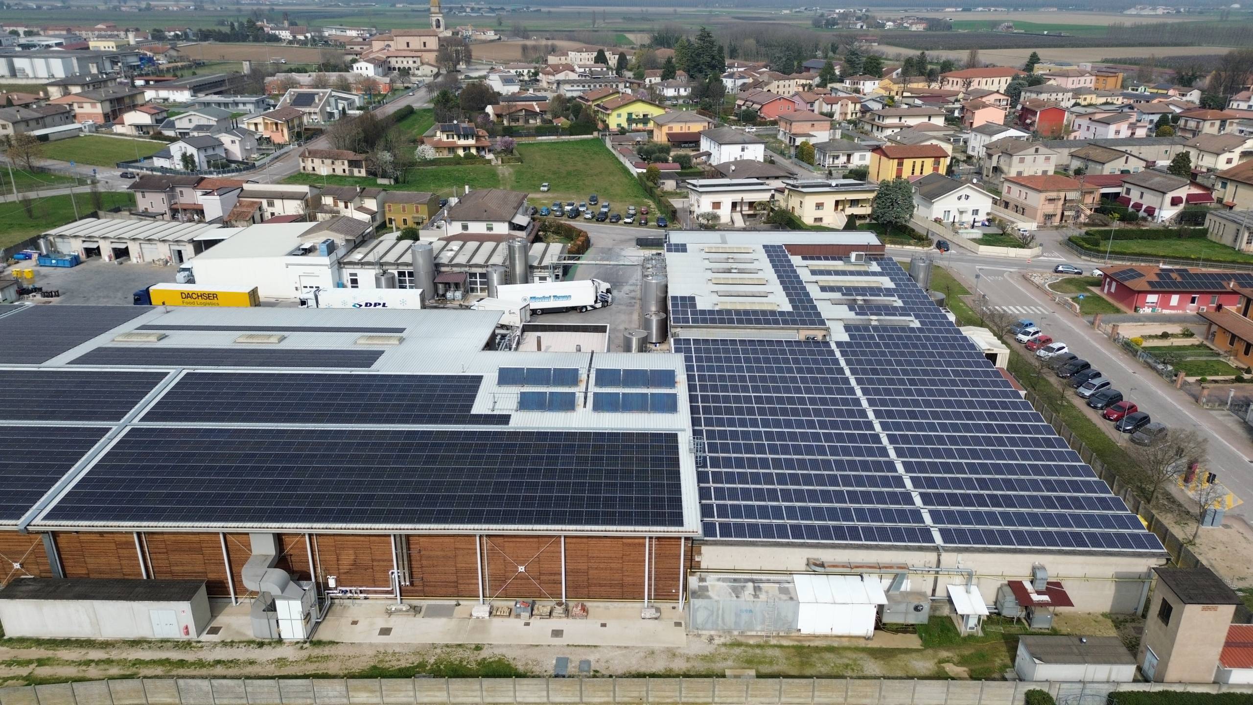 DCIM100MEDIADJI_0459JPG - Pleion Aerial view of an industrial facility with large solar panels covering the roof, loading bays and delivery trucks nearby, and a residential town beyond.