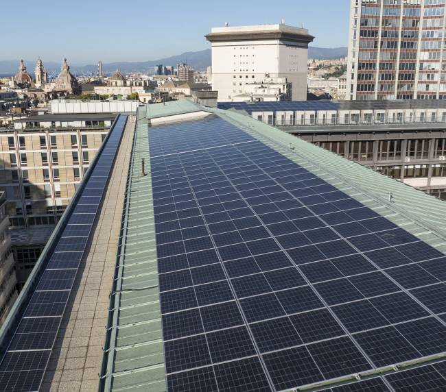Rooftop covered in dark solar panels overlooking a dense city with domed towers and distant hills/ocean.