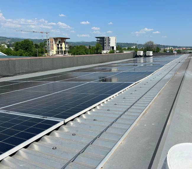 Rooftop solar panels in rows with a city and green hills beyond under a clear blue sky.”] ,