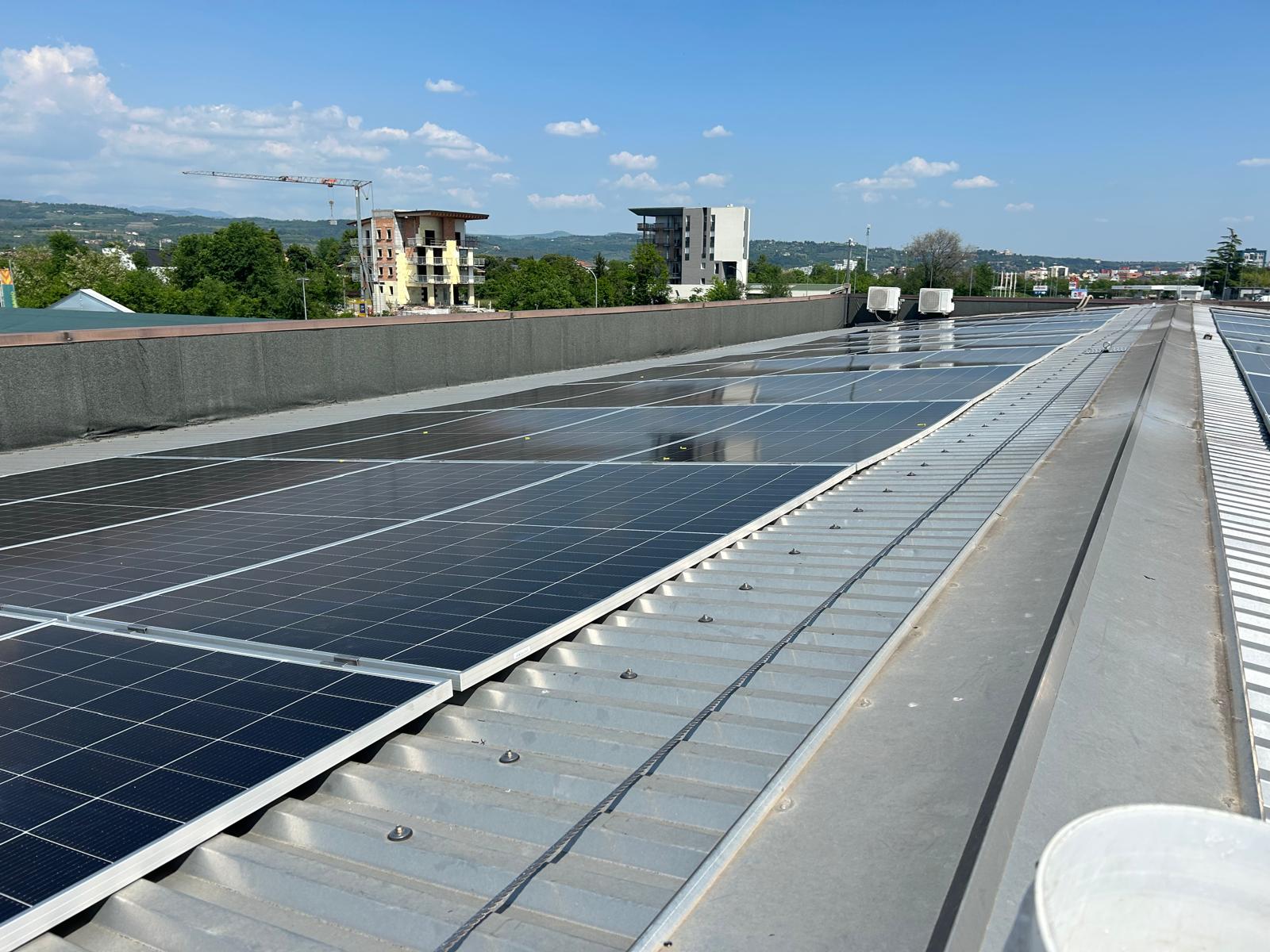 Rooftop solar panels in rows with a city and green hills beyond under a clear blue sky.”] ,