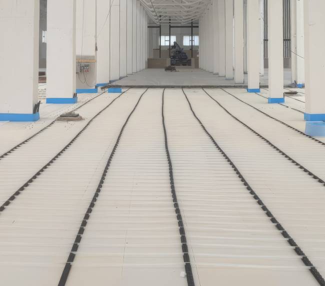 Interior of a large unfinished building with white columns and blue base trim, showing radiant floor heating pipes laid in long parallel loops across the floor.
