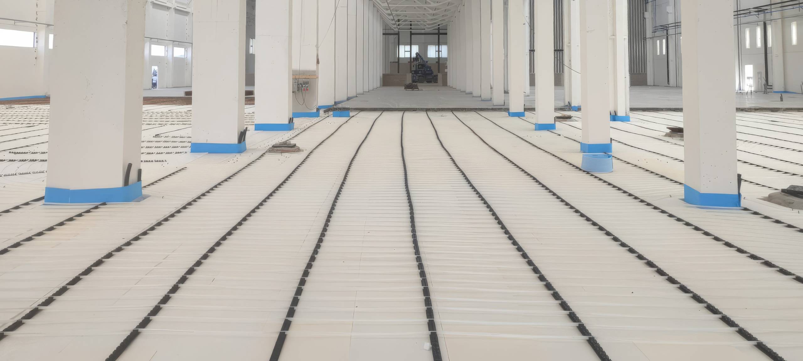 fr6-tz-upscale-2x - Pleion Interior of a large unfinished building with white columns and blue base trim, showing radiant floor heating pipes laid in long parallel loops across the floor.