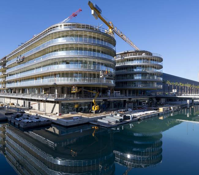 Curved glass-fronted building under construction at the waterfront with a large yellow crane overhead.