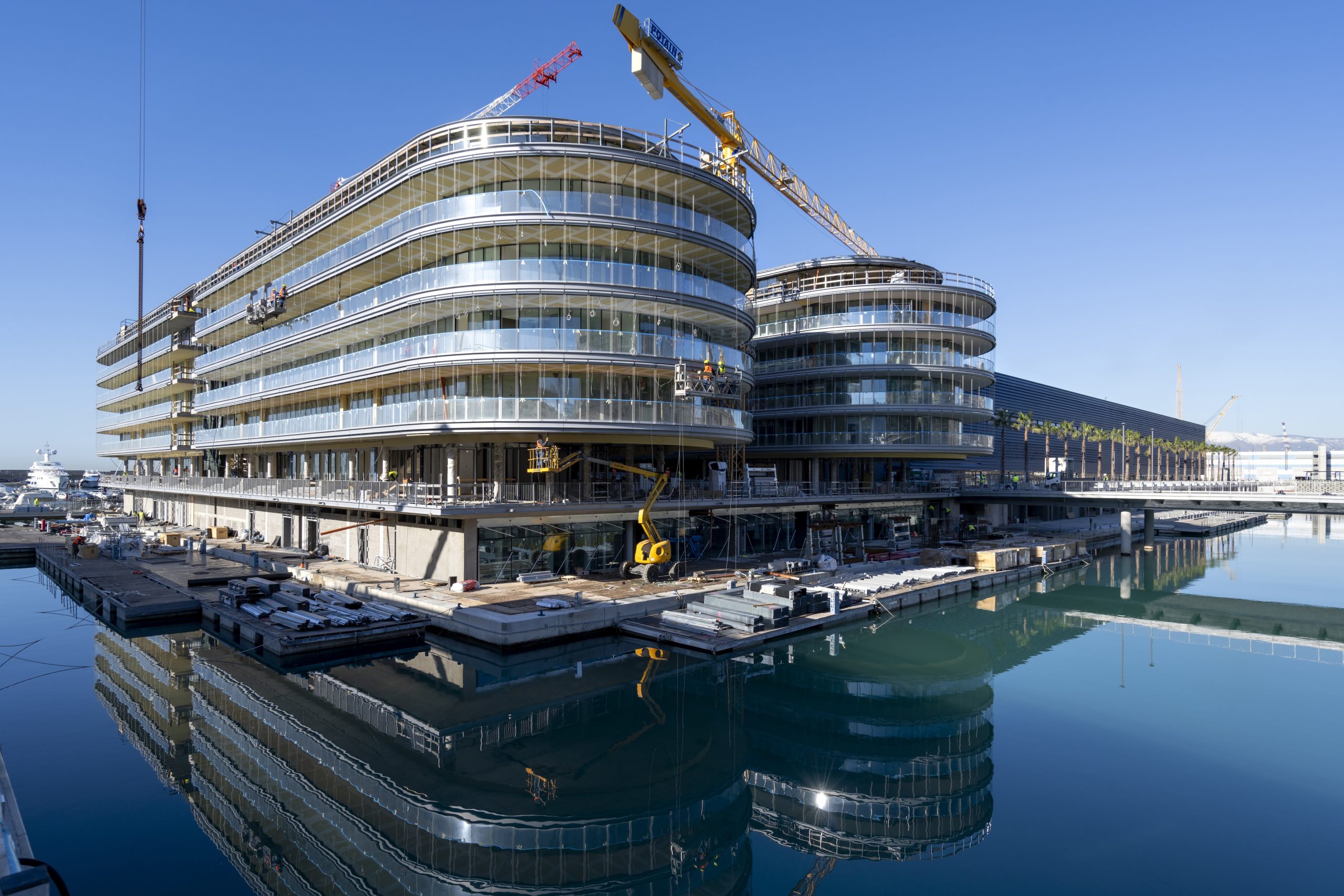 ©Resstende_Waterfront_Dallas-110_Ph-Andrea-Botto-1-2-scaled - Pleion Curved glass-fronted building under construction at the waterfront with a large yellow crane overhead.