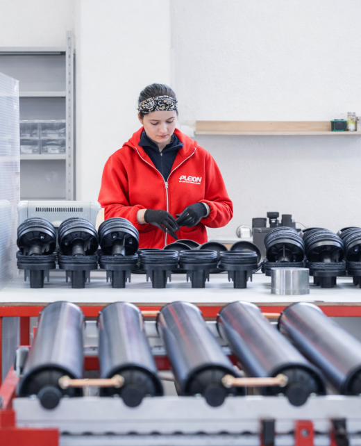 Worker in red jacket assembles black plastic components on a workshop table with rack of parts nearby.